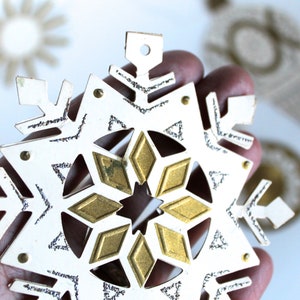 May include: A white paper snowflake ornament with a gold glitter design. The snowflake has a hole at the top for hanging.