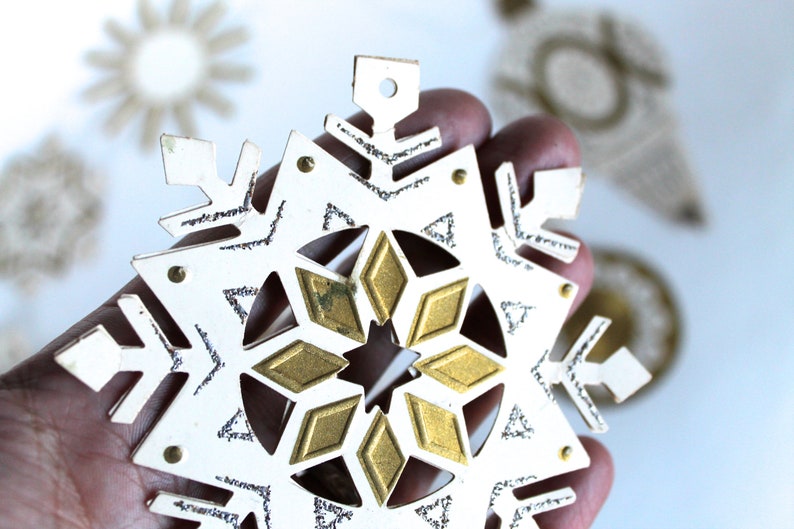 May include: A white paper snowflake ornament with a gold glitter design. The snowflake has a hole at the top for hanging.