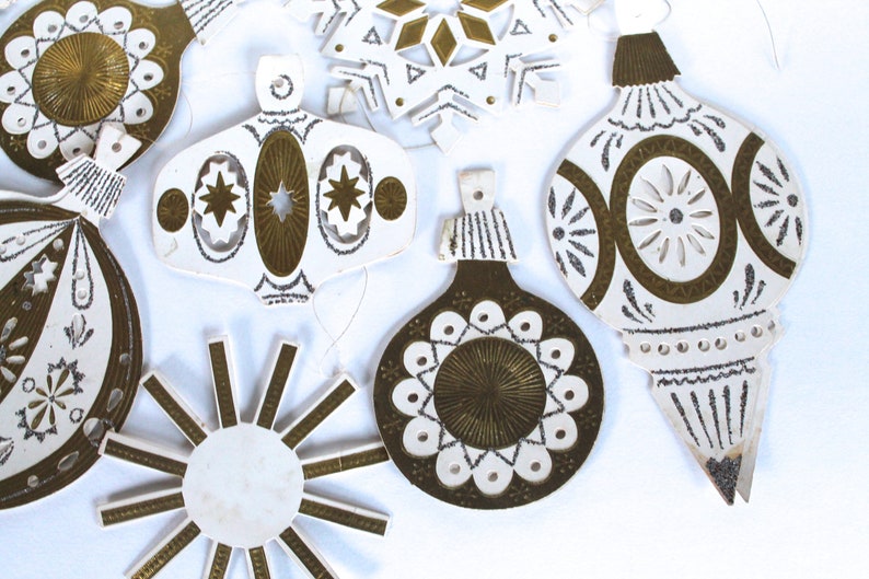 May include: A set of eight vintage paper Christmas ornaments with gold and white designs. The ornaments are shaped like stars, snowflakes, and balls.