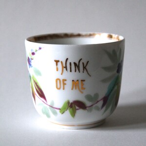May include: A white porcelain teacup with a gold rim and floral design. The cup has the words "Think of Me" written in gold lettering.