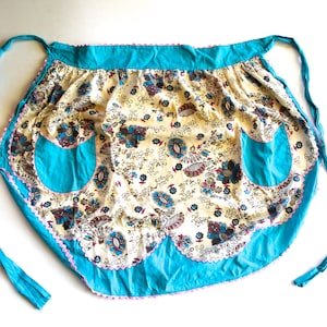 May include: A turquoise and white floral print apron with two pockets. The apron has a scalloped edge and pink trim.