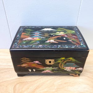 May include: Black wooden jewelry box with a hinged lid. The lid is decorated with a painted scene of a Japanese garden with a mountain, trees, and a pagoda. The box has a brass keyhole and sits on four small feet.