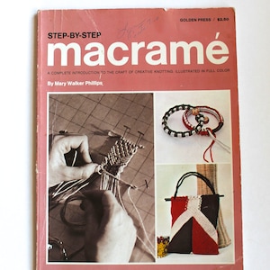 May include: A book titled "Step-by-Step Macrame" with a pink cover and a photo of a person making a macrame knot. The book is a complete introduction to the craft of creative knotting, illustrated in full color. The book is by Mary Walker Phillips.