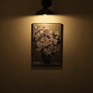 May include: A framed floral painting featuring a bouquet of white flowers in a dark green vase. The artwork is illuminated by a spotlight from a black, industrial-style fixture mounted above. The background is a warm, neutral wall.