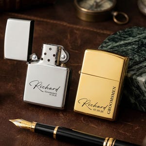 Puede incluir: Dos encendedores personalizados, uno plateado y otro dorado, descansan sobre una superficie de cuero. El encendedor plateado est&aacute; abierto, revelando la inscripci&oacute;n "Richard" y una fecha. El encendedor dorado est&aacute; cerrado, con la inscripci&oacute;n "Richard", "Groomsmen" y una fecha.