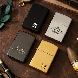 Puede incluir: Cuatro encendedores personalizados en negro, plateado y dorado, con grabados personalizados. Un encendedor negro presenta la letra "A", otro plateado tiene un dise&ntilde;o de monta&ntilde;a y el nombre "Zachary". Un tercer encendedor negro est&aacute; grabado con "Justin Groomman", y el &uacute;ltimo es dorado con la letra "M".