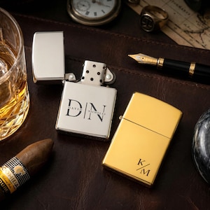 Puede incluir: Dos encendedores personalizados, uno plateado y otro dorado, descansan sobre una superficie de cuero. El encendedor plateado est&aacute; abierto, mostrando las iniciales "DN" y el nombre "David Nicholas". El encendedor dorado muestra las iniciales "K/M". Tambi&eacute;n se ven un vaso de l&iacute;quido &aacute;mbar, un cigarro y un bol&iacute;grafo.
