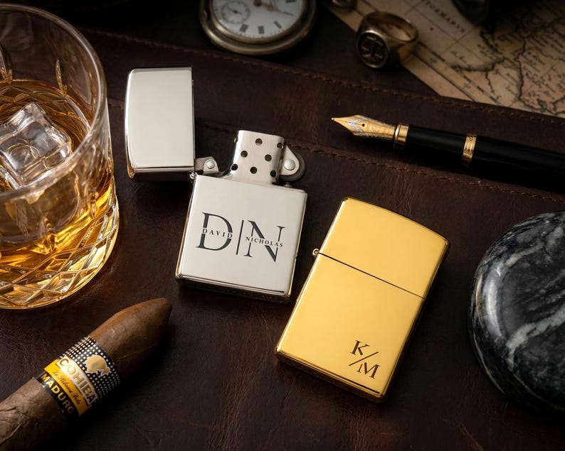 Puede incluir: Dos encendedores personalizados, uno plateado y otro dorado, descansan sobre una superficie de cuero. El encendedor plateado est&aacute; abierto, mostrando las iniciales "DN" y el nombre "David Nicholas". El encendedor dorado muestra las iniciales "K/M". Tambi&eacute;n se ven un vaso de l&iacute;quido &aacute;mbar, un cigarro y un bol&iacute;grafo.