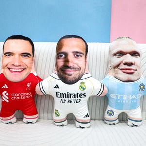 Peut inclure: Trois poupées en peluche avec des visages imprimés, chacune portant un uniforme de football. L'une porte un maillot rouge de Liverpool, une autre un maillot blanc du Real Madrid et la troisième un uniforme bleu clair de Manchester City.