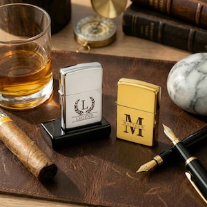 Puede incluir: Dos encendedores met&aacute;licos personalizados, uno plateado y otro dorado, se exhiben sobre una superficie de cuero marr&oacute;n. El encendedor plateado tiene la palabra "LEGEND" y un dise&ntilde;o de corona de laurel. El encendedor dorado tiene el nombre "MATTHEW".