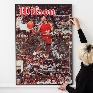 Peut inclure: Affiche encadrée représentant Michael Jordan en uniforme de basket-ball rouge, en l'air, avec un ballon. L'affiche porte le mot "Wilson" en haut et la signature de Michael Jordan en bas. L'arrière-plan montre un stade bondé.