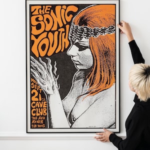 May include: A framed poster with a black border featuring a retro design. The poster has orange and black text that reads "THE SONIC YOUTH" and details for a concert at the Cave Club. The artwork includes a stylized illustration of a woman with orange hair.