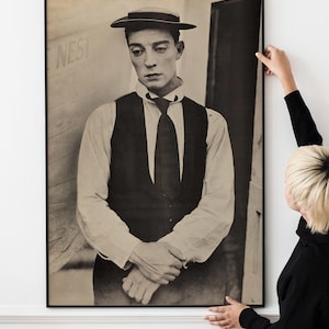 Puede incluir: Impresión enmarcada en blanco y negro que presenta un retrato de Buster Keaton. La imagen muestra a Keaton con traje, corbata y sombrero, con la palabra "NEST" visible en el fondo. La impresión está siendo sostenida por una persona.