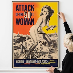 May include: A framed movie poster for "Attack of the 50 Foot Woman." The poster features a giant woman towering over a city, holding a car. The title is in large, bold letters, with the names of the actors and production credits below.