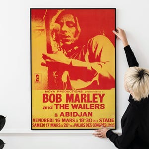 May include: A framed poster with a red and yellow color scheme featuring Bob Marley and The Wailers. The poster includes the text "BOB MARLEY and THE WAILERS à ABIDJAN" and concert details. The image has a vintage aesthetic.