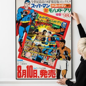 以下が含まれることがあります： スーパーマン対モハメド・アリのフレーム入りポスター。アートワークは、ボクシングの姿勢のスーパーマンとボクシングショーツのアリを描いています。日本語のテキストと日付「8月10日、発売」が表示されています。ポスターは人が持っています。