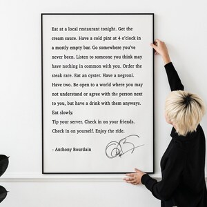 Puede incluir: Un estampado en blanco y negro enmarcado con texto de Anthony Bourdain. El texto incluye frases como "Come en un restaurante local esta noche" y "Disfruta del viaje". Una persona sostiene el marco contra una pared blanca.