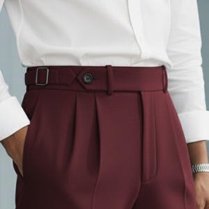 May include: Burgundy-colored dress pants with a high waist and a matching belt. The pants have a button closure and a metal buckle. The pleats and tailored design suggest a formal or business style.