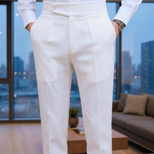 Könnte beinhalten: Weiße Hose mit hohem Bund und Knopfverschluss. Die Hose hat eine maßgeschneiderte Passform und wird mit einem weißen Hemd kombiniert. Das Bild wurde in Innenräumen aufgenommen, mit Blick auf die Skyline durch ein großes Fenster.