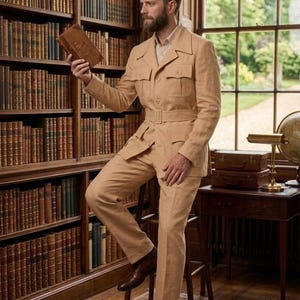 May include: A tan safari suit with a belted jacket and matching trousers. The suit is worn with brown leather shoes. The person is holding a book in a library setting with bookshelves and a window.