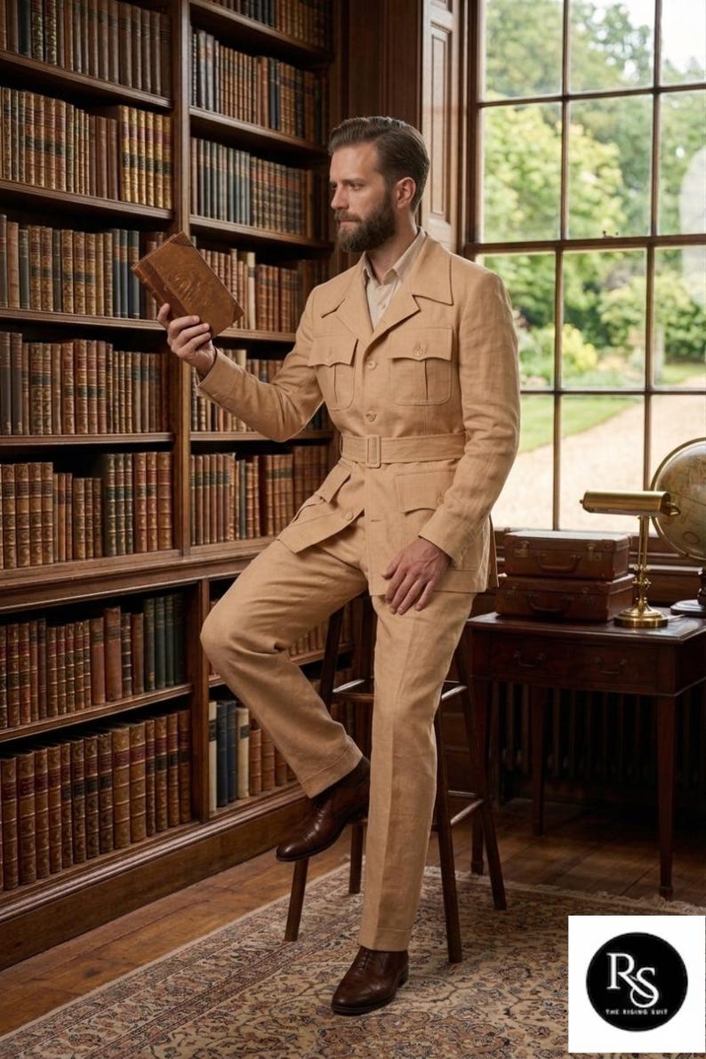 May include: A tan safari suit with a belted jacket and matching trousers. The suit is worn with brown leather shoes. The person is holding a book in a library setting with bookshelves and a window.