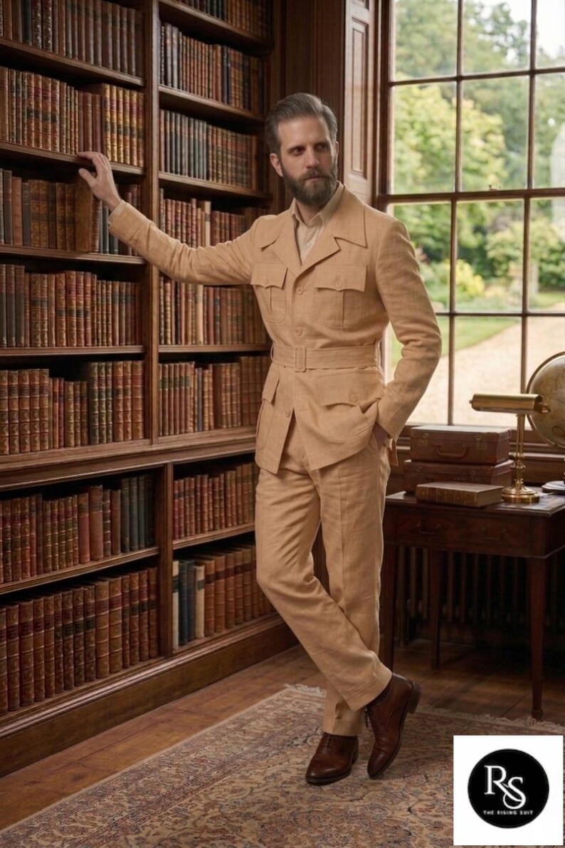 May include: A tan safari suit with a belted waist and multiple pockets. The suit is worn with a light-colored shirt and brown leather shoes. The person is standing in a library with bookshelves filled with books.