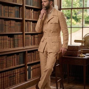 May include: A tan linen suit with a belted jacket and matching trousers. The suit has multiple pockets and is paired with brown leather shoes. The setting is a library with a large bookshelf and a wooden desk.