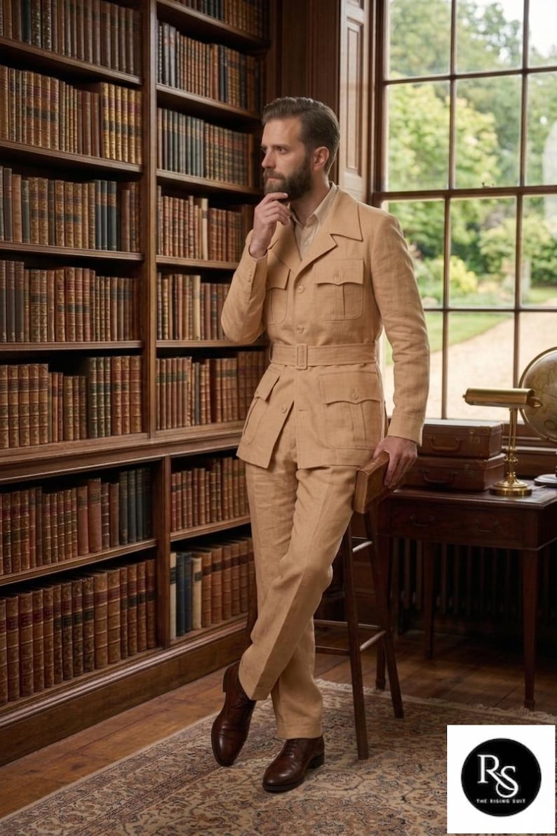 May include: A tan linen suit with a belted jacket and matching trousers. The suit has multiple pockets and is paired with brown leather shoes. The setting is a library with a large bookshelf and a wooden desk.