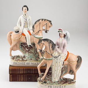 May include: Two porcelain figurines of a Prince and Princess on horseback. The Prince wears a white suit and the Princess a pink dress. The horses are tan with black manes. The figurines sit on bases with the words "Prince of Wales" and "Princess".