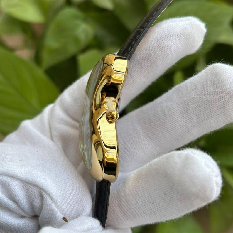 May include: A gold-toned wristwatch with a black leather band. The watch has a round face and a small crown. The watch is shown from the side, highlighting its polished finish and elegant design. The watch is a classic accessory.
