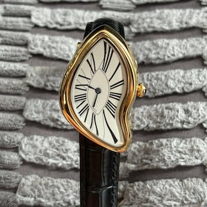 May include: A gold-toned wristwatch with a distorted, melting design. The watch face is white with black Roman numerals and hands. The black leather band complements the watch's unique, surreal aesthetic. The watch is a luxury item.