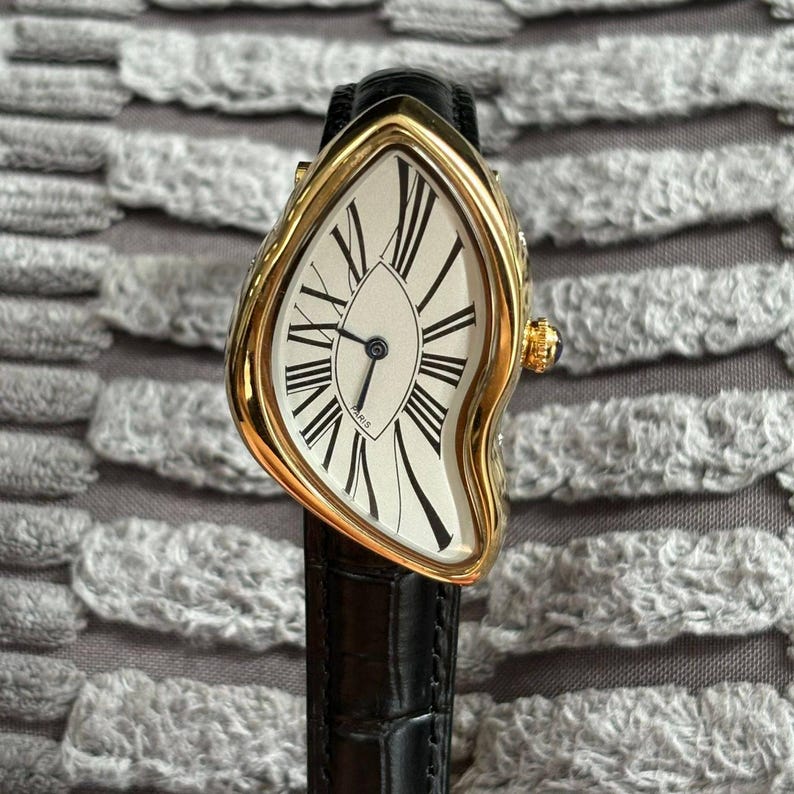May include: A gold-toned wristwatch with a distorted, melting design. The watch face is white with black Roman numerals and hands. The black leather band complements the watch's unique, surreal aesthetic. The watch is a luxury item.
