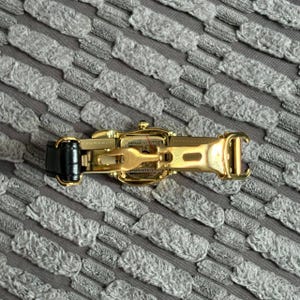 May include: A gold-toned watch with a black leather band is displayed open, revealing the clasp mechanism. The watch is set against a textured gray background, highlighting its intricate design and craftsmanship. The watch's details are clearly visible.