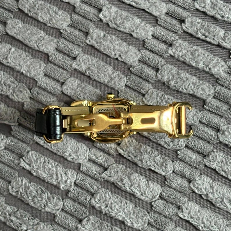 May include: A gold-toned watch with a black leather band is displayed open, revealing the clasp mechanism. The watch is set against a textured gray background, highlighting its intricate design and craftsmanship. The watch's details are clearly visible.