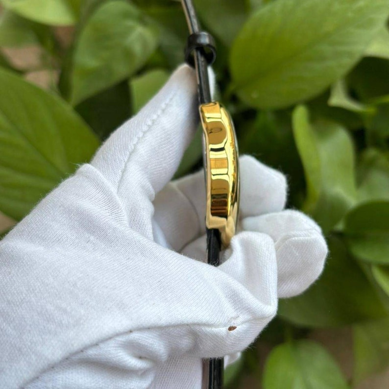 May include: A gold-toned wristwatch with a black leather band. The watch face is not visible, but the side profile shows a rounded, rectangular shape. The watch is held by a gloved hand against a backdrop of green foliage.