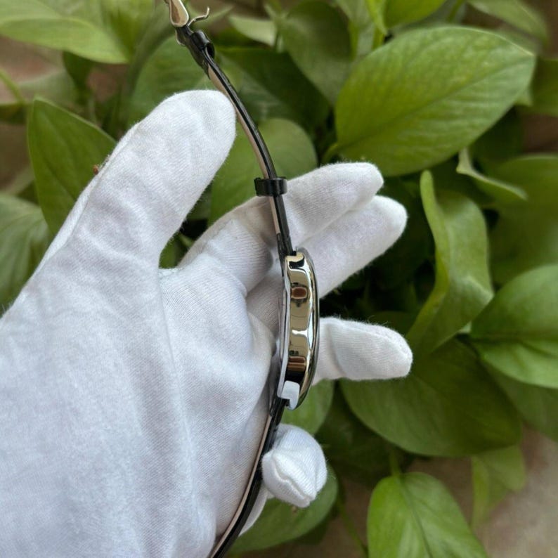 Pode incluir: Um rel&oacute;gio de pulso prateado com uma pulseira de couro preta. O rel&oacute;gio tem um mostrador redondo com um mostrador preto e ponteiros prateados. O rel&oacute;gio &eacute; segurado por uma m&atilde;o enluvada contra um fundo de folhagem verde.