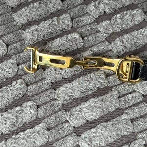 May include: Close-up of a gold-toned metal belt buckle with a black leather strap. The buckle features a hook and a decorative design. The background is a textured gray fabric.