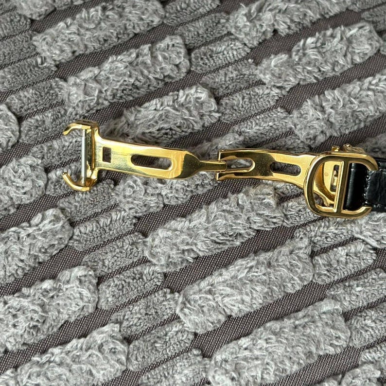 May include: Close-up of a gold-toned metal belt buckle with a black leather strap. The buckle features a hook and a decorative design. The background is a textured gray fabric.
