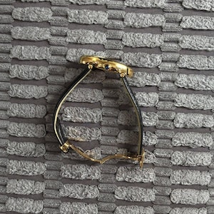 May include: A gold-toned wristwatch with a black leather band. The watch face is round with a blue gem. The watch is displayed on a textured gray surface.