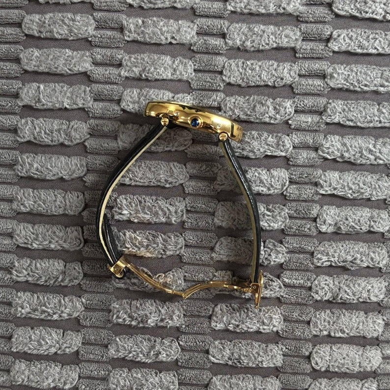 May include: A gold-toned wristwatch with a black leather band. The watch face is round with a blue gem. The watch is displayed on a textured gray surface.