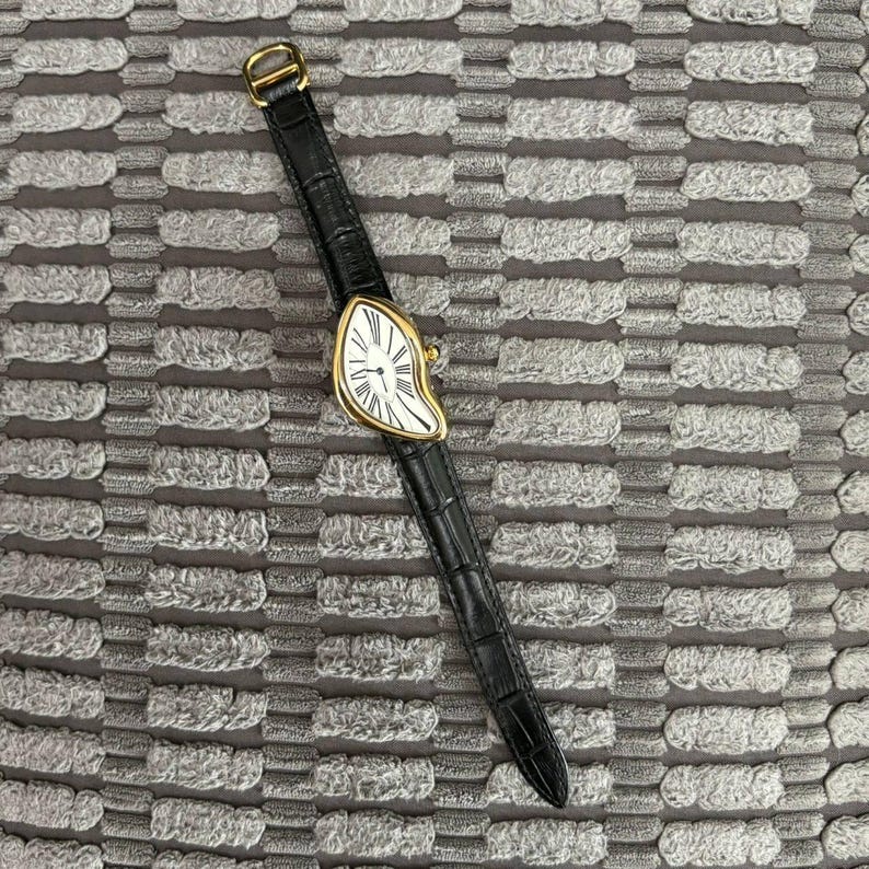 Gold-tone surreal melting watch with a distorted dial and black leather strap, laid flat on a textured gray fabric background. The dial features Roman numerals and a blue-tinted hand, evoking Salvador Dali’s melting clock style. Ideal for showcasing the artistic and bold design of the watch.