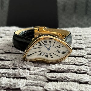 May include: A gold-toned wristwatch with a unique, eye-shaped face featuring Roman numerals and blue hands. The watch has a black leather band and a gold clasp. The watch is displayed on a textured gray surface.
