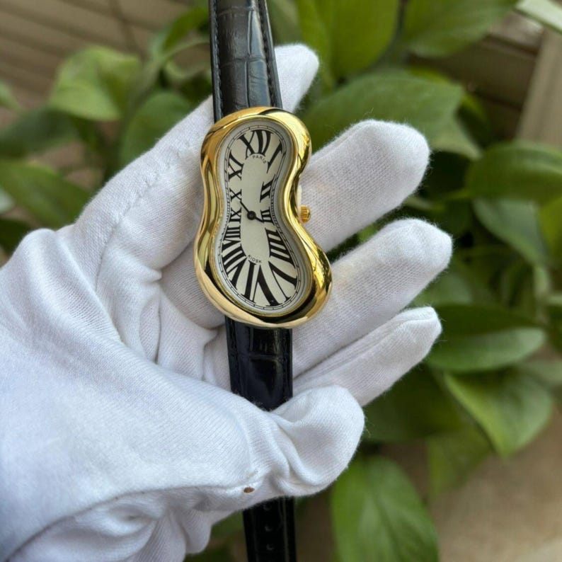 🕰️ Surrealist melting dial watch held in a white gloved hand, featuring a gold-toned curved case, black Roman numerals, and a black leather strap, photographed outdoors with green leaves in the background.