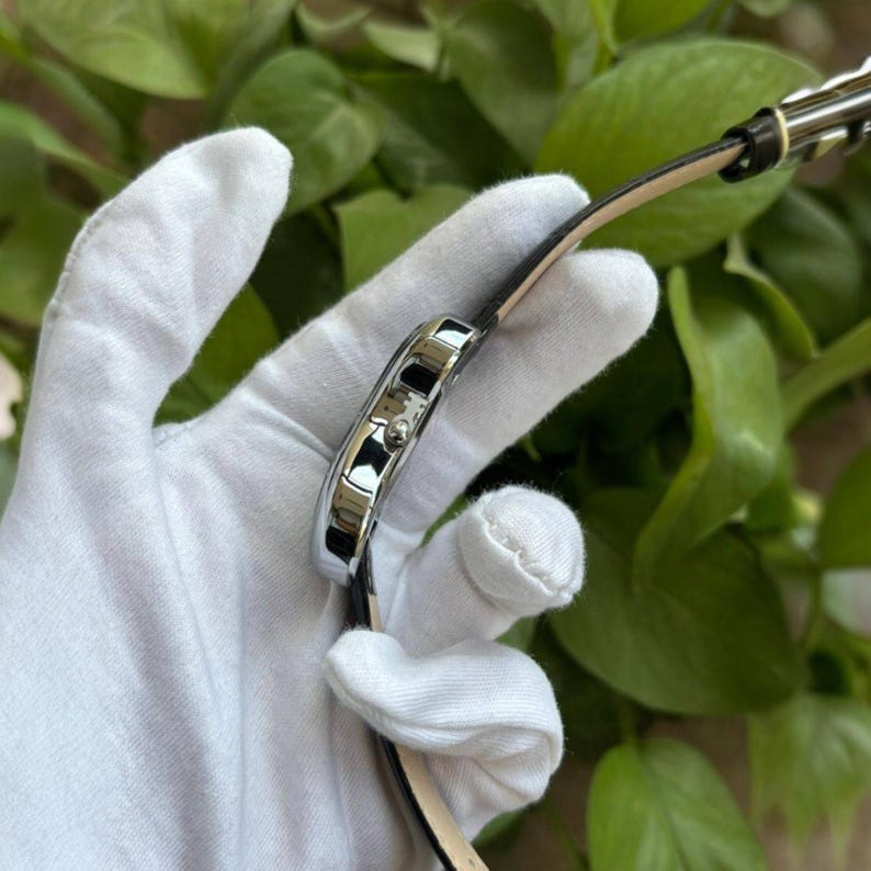 Pode incluir: Um rel&oacute;gio de pulso retangular prateado com uma pulseira de couro preta. O mostrador do rel&oacute;gio &eacute; vis&iacute;vel e a pulseira est&aacute; presa &agrave; caixa do rel&oacute;gio. O rel&oacute;gio &eacute; segurado por uma m&atilde;o enluvada em um fundo de folhagem verde.