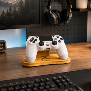 Può includere: Un controller per videogiochi bianco poggia su un supporto in legno chiaro. Il supporto presenta una grafica di montagna e il testo B.Springer stampato sulla parte anteriore. Il controller ha levette analogiche nere, un touchpad nero e tasti.