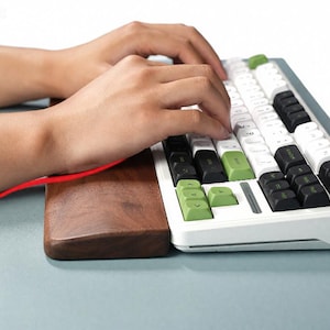 Könnte beinhalten: Eine mechanische Tastatur mit weißen, schwarzen und grünen Tastenkappen. Die Tastatur hat einen weißen Rahmen und eine Handgelenkauflage aus Holz. Die Tasten werden von den Händen einer Person gedrückt. Die Tastatur liegt auf einer hellblauen Oberfläche.