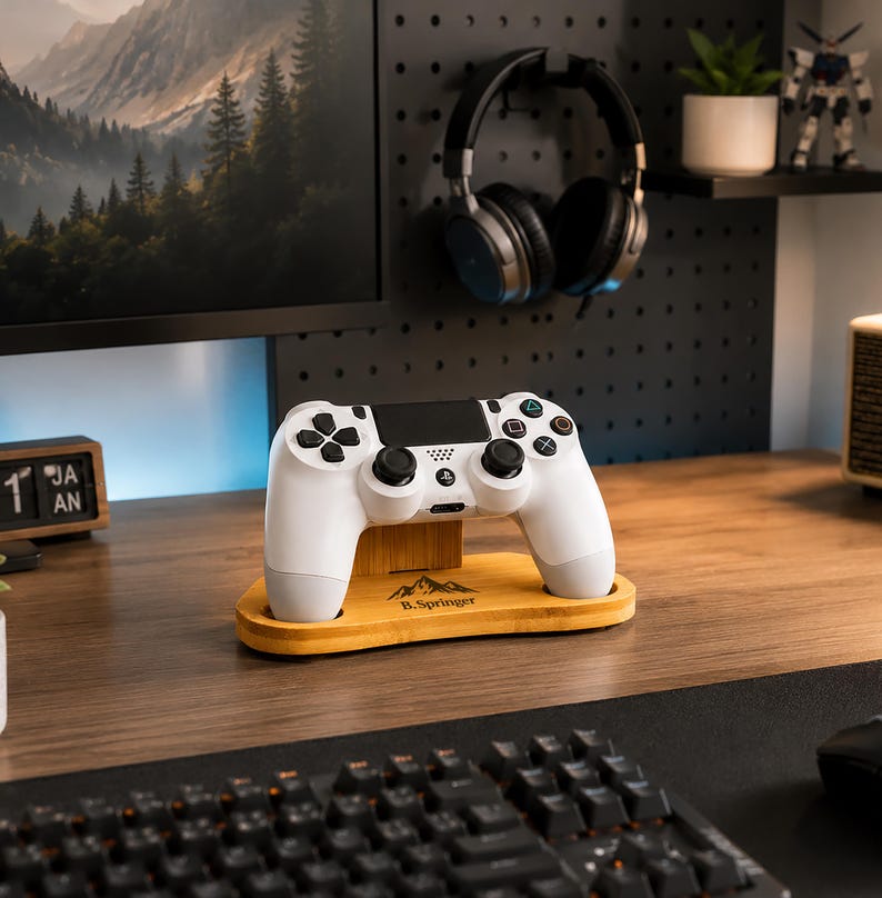 May include: A white video game controller rests on a light-colored wooden stand. The stand features a mountain graphic and the text B.Springer printed on the front. The controller has black analog sticks, a black touchpad, and standard buttons.