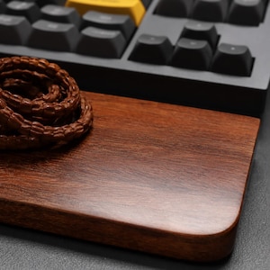 May include: A rectangular wooden wrist rest with a dark brown grain pattern, positioned in front of a black mechanical keyboard featuring dark grey keys and a single yellow key. A coiled brown beaded bracelet rests on the left side of the wood.