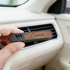 Könnte beinhalten: Ein Auto-Luftauslass-Clip mit einem dunkelgrauen Gehäuse und einem Holzakzent. Der Holzakzent ist mit dem Namen "Luna" graviert. Der Clip ist für die Befestigung an einer Auto-Luftauslassöffnung konzipiert.