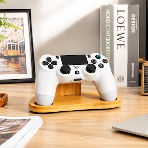 May include: A white wireless game controller rests on a light-colored wooden stand. The controller features black analog sticks, a central touchpad, and standard action buttons including a triangle, circle, cross, and square in their respective colors.